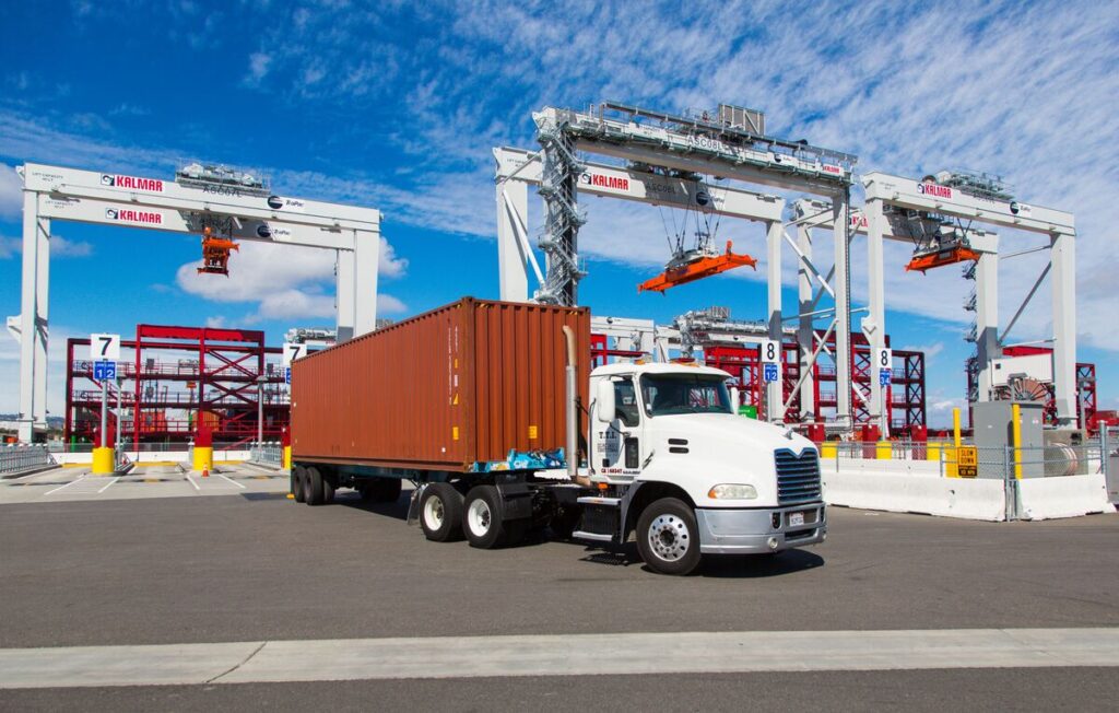 "A white truck with a large orange shipping container is parked in front of several large Kalmar container cranes at a shipping yard.