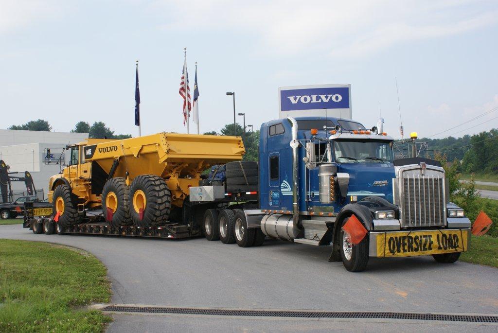 A white semi-truck labeled '49082' with the 'Albee' logo transports a large Vermeer industrial machine on a flatbed trailer. The machine has large tires and a yellow frame, indicating heavy-duty equipment. The truck has an 'Oversize Load' sign on the front bumper.