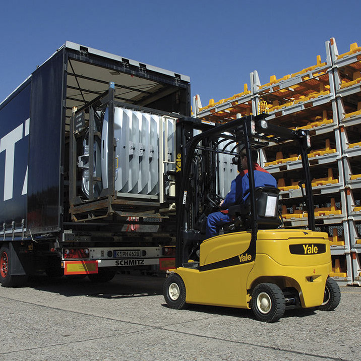 "A forklift operator is using a yellow Yale forklift to load or unload a large metal frame from a truck. The truck has an open back, revealing the cargo inside. The truck has a license plate with the number 'K PH 4620' and the brand 'SCHMITZ' visible. In the background, there are multiple stacks of metal racks with yellow components stored on them. The scene takes place outdoors on a concrete surface under a clear blue sky.