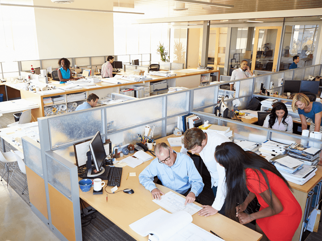 An open office space with multiple employees working at their desks and collaborating in groups. The workspace is bright and modern, featuring cubicles, computers, and office supplies, with large windows letting in natural light