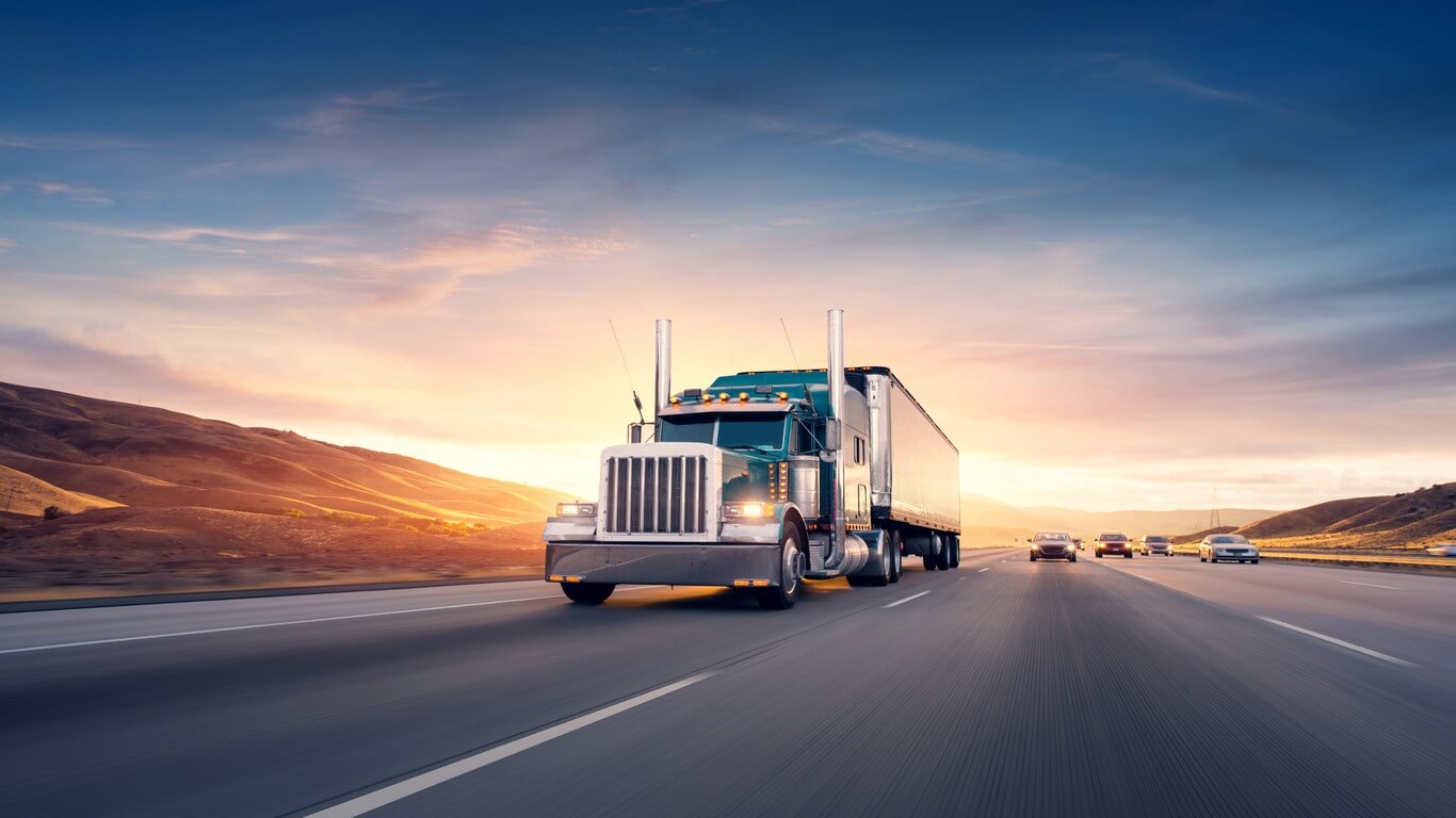 A semi-truck driving on a highway at sunset with rolling hills in the background and several cars following behind.